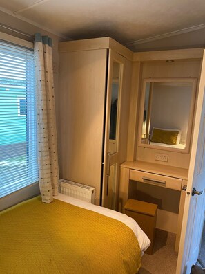 A compact bedroom with a bed, a bedside table, a mirror, a radiator, and a window with curtains. - Cosy Grasmere Static Caravan (Natland)