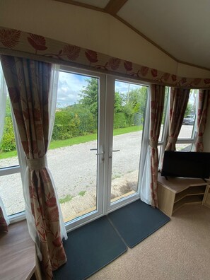 A room with a sliding glass door leading to a gravel driveway, a small TV on a wooden stand, and a carpeted floor. - Cosy Rydal Static Caravan (Natland)