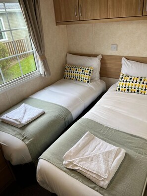 A room with two single beds, each with a pillow and a folded towel. There is a window with curtains, and a wooden cabinet above the beds. - Cosy Rydal Static Caravan (Natland)