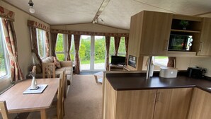 A spacious kitchen with wooden cabinets, a dining area with a table and chairs, and large windows with curtains. - Cosy Rydal Static Caravan (Natland)