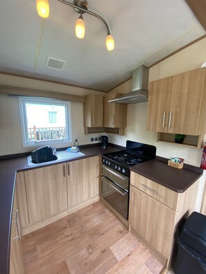 A compact kitchen with wooden cabinets, a gas stove, an oven, and a window with a view of a balcony. - Cosy Rydal Static Caravan (Natland)