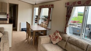 A cozy living area with a sofa, a dining table, and chairs. There is a kitchenette with cabinets and a radiator. The room has large windows with curtains. - Cosy Rydal Static Caravan (Natland)