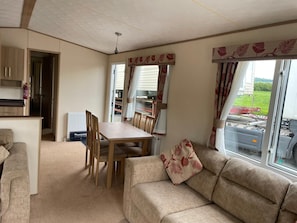 A cozy living area with a sofa, a dining table, and chairs. There is a kitchenette with cabinets and a radiator. The room has large windows with curtains. - Cosy Rydal Static Caravan (Natland)
