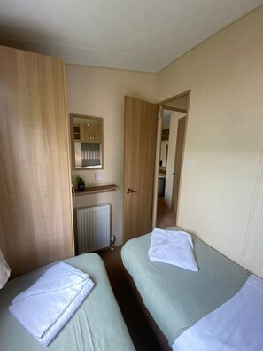 A small, neatly arranged room with two single beds, a wooden wardrobe, a radiator, and a mirror on the wall. - Cosy Rydal Static Caravan (Natland)