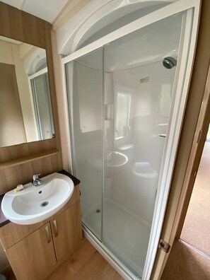 A bathroom with a glass-enclosed shower, a sink with a mirror above it, and wooden cabinets. - Cosy Rydal Static Caravan (Natland)