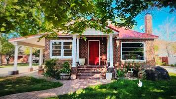 A brick house with a red door, a covered porch, and a front yard with potted plants and a large rock.
