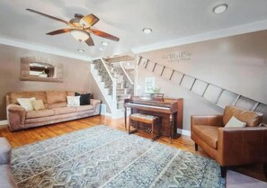 Ein geräumiges Wohnzimmer mit Holzboden, Deckenventilator, Klavier, Treppe und einer Couch.