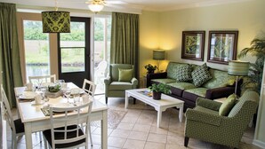 A well-lit living room with a dining area, green patterned furniture, and a view through the glass door.