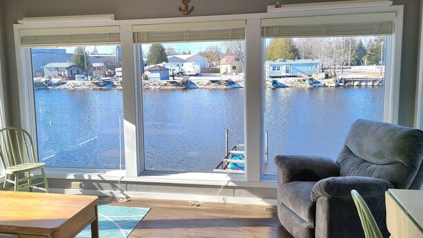 Een kamer met grote ramen met uitzicht op een jachthaven, een grijze fauteuil, een houten tafel en een groene stoel.