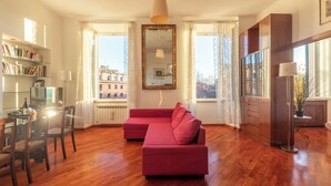 Un salon spacieux avec un canapé rouge, un plancher en bois, un coin repas comprenant des chaises et une table, et de grandes fenêtres offrant une vue sur les bâtiments environnants.