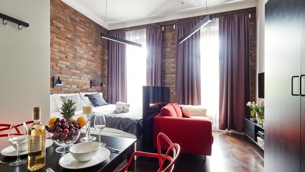 Une chambre d’hôtel moderne avec un mur en briques, un canapé rouge, une table à manger avec une bouteille de vin et un lit donnant sur la ville.