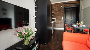 Un salon moderne avec un téléviseur à écran plat, un meuble de rangement noir, un vase de tulipes roses et un canapé orange vif.