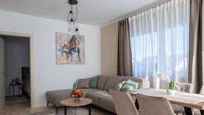 Una sala de estar moderna con sofá, mesa de centro y zona de comedor. Hay un cuadro en la pared y una ventana con cortinas.