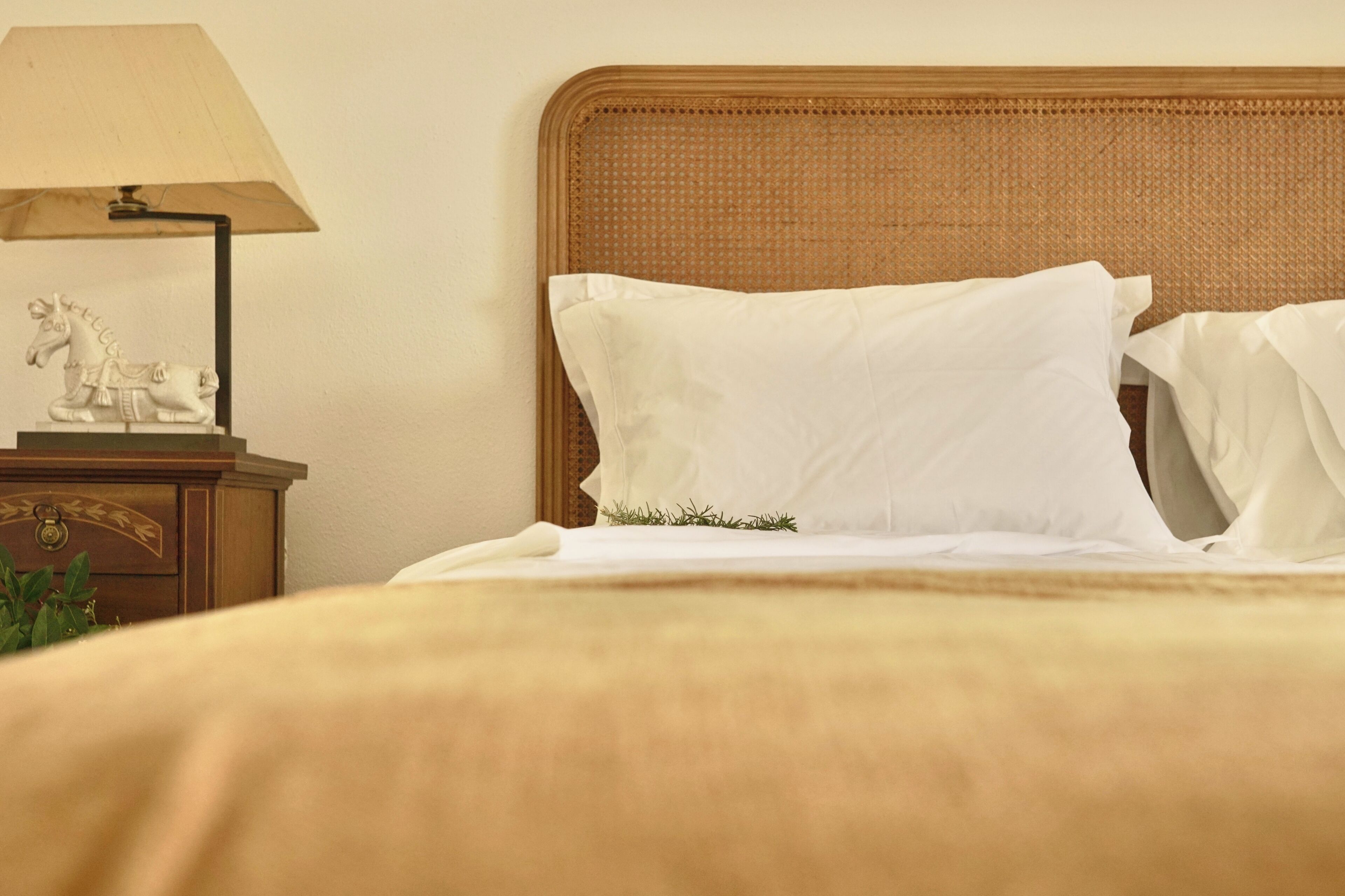A neatly made bed with white linens and a decorative pillow, a bedside table with a lamp and a small sculpture, and a wicker headboard.