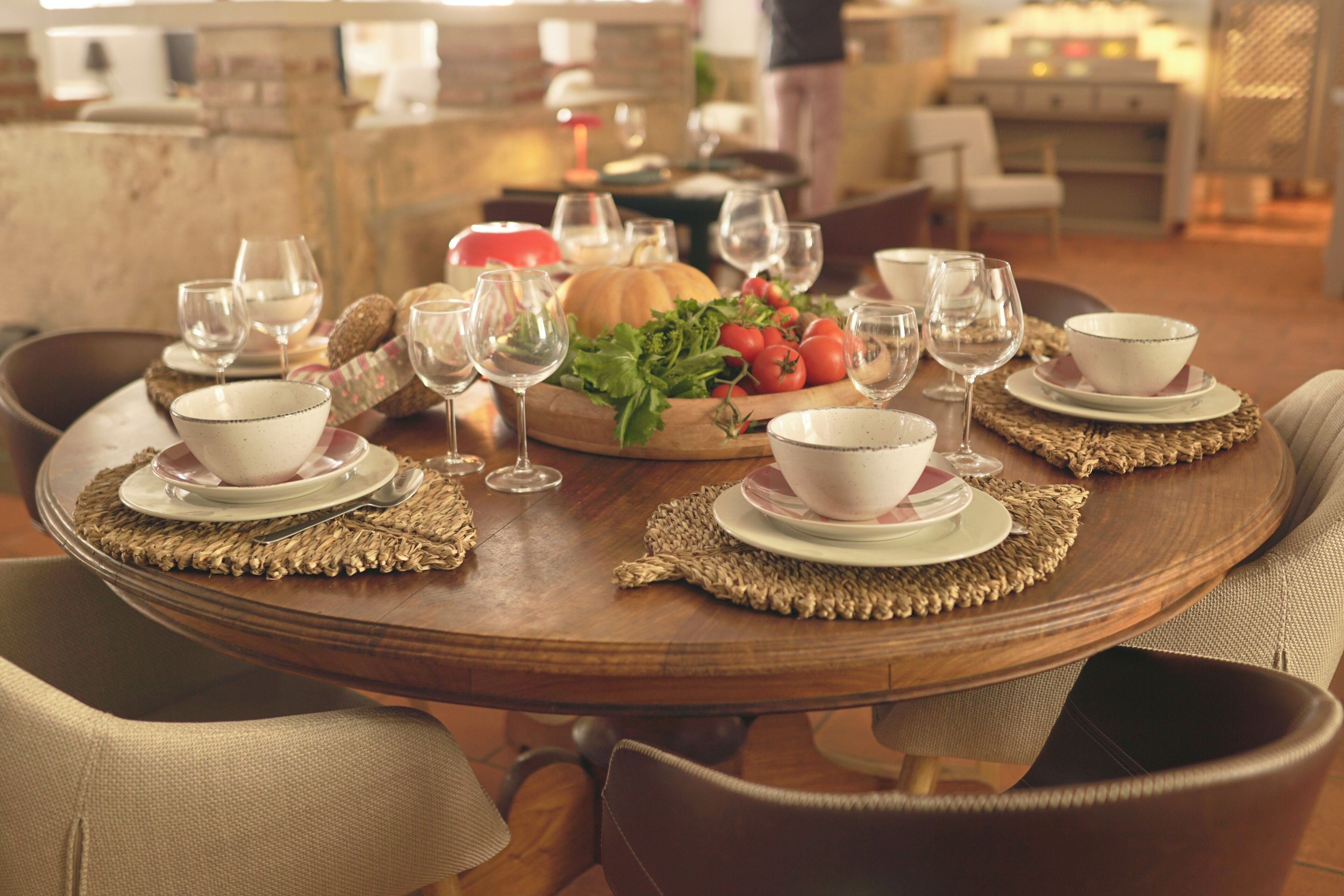 A round dining table set with white bowls, wine glasses, and a centerpiece of vegetables.