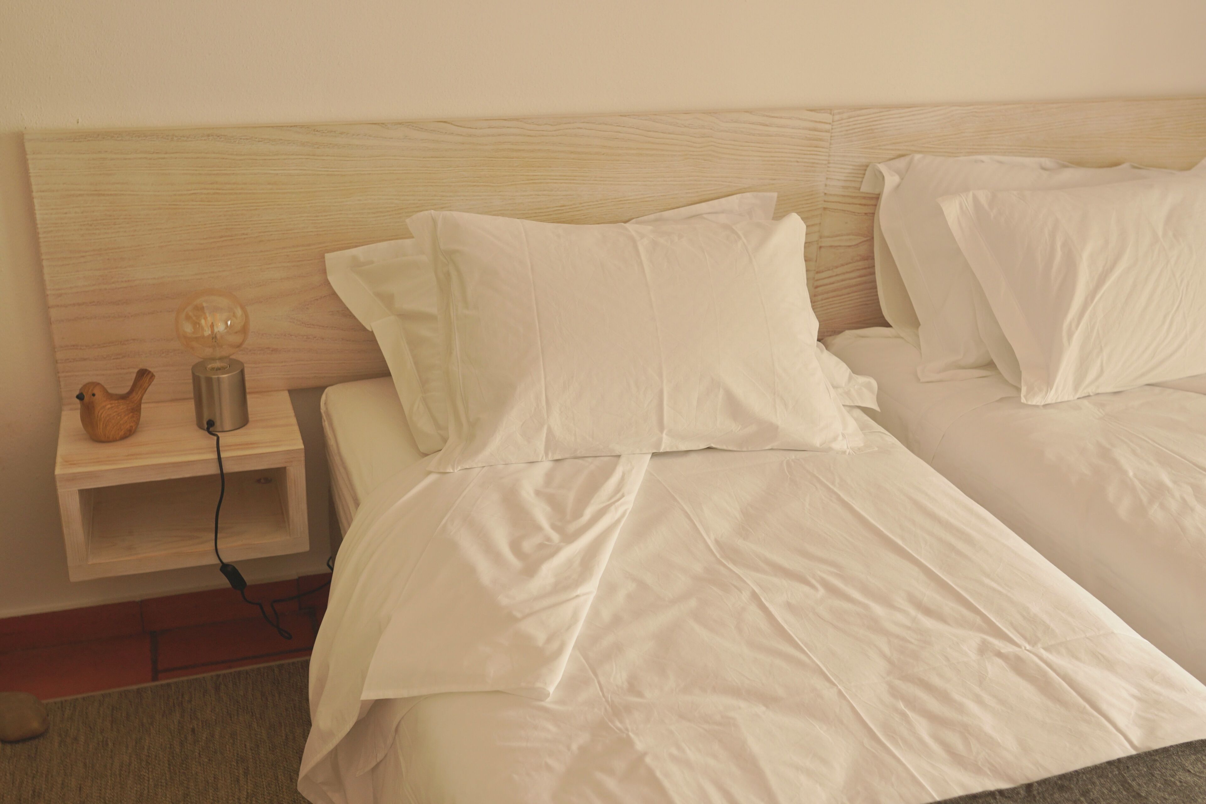 Una cama bien hecha con sábanas blancas y cabecera de madera. Se observa una mesita de noche con una lámpara y un pequeño objeto decorativo.