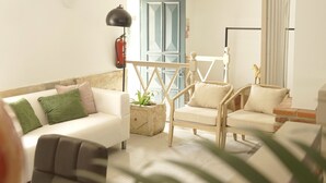 Una sala de estar con un sofá blanco, una silla de madera y una mesita. Hay una planta en el suelo y una lámpara sobre un pedestal.