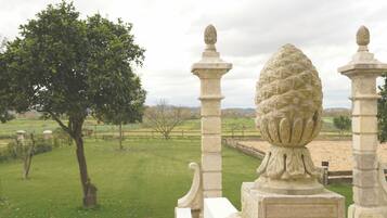 Een stenen balustrade met een decoratieve vaas als middelpunt, een grasveld en een boom op de achtergrond.