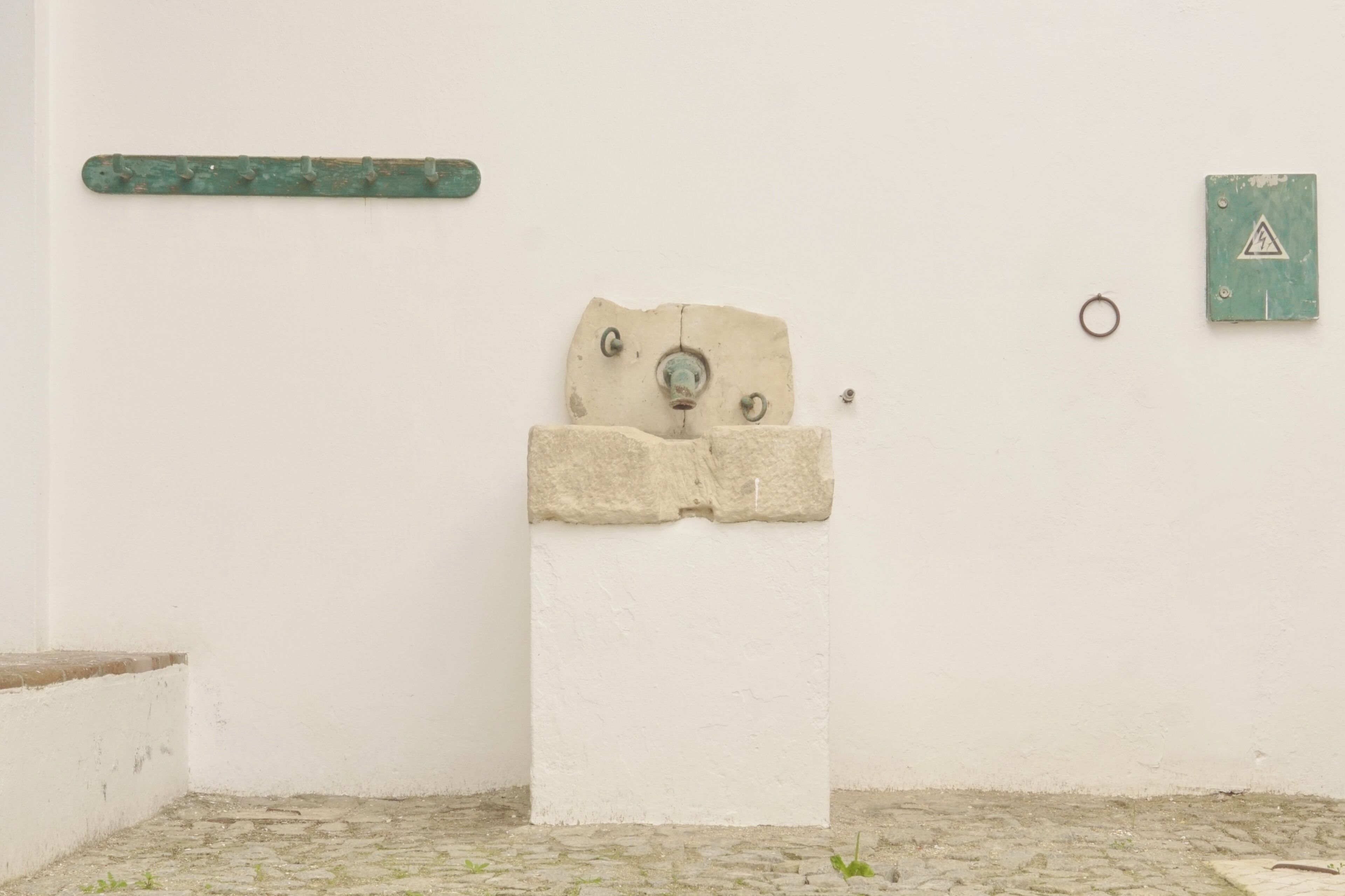 Estructura de piedra con una abertura circular, posiblemente un pozo, frente a una pared blanca con un letrero verde y un aro metálico.
