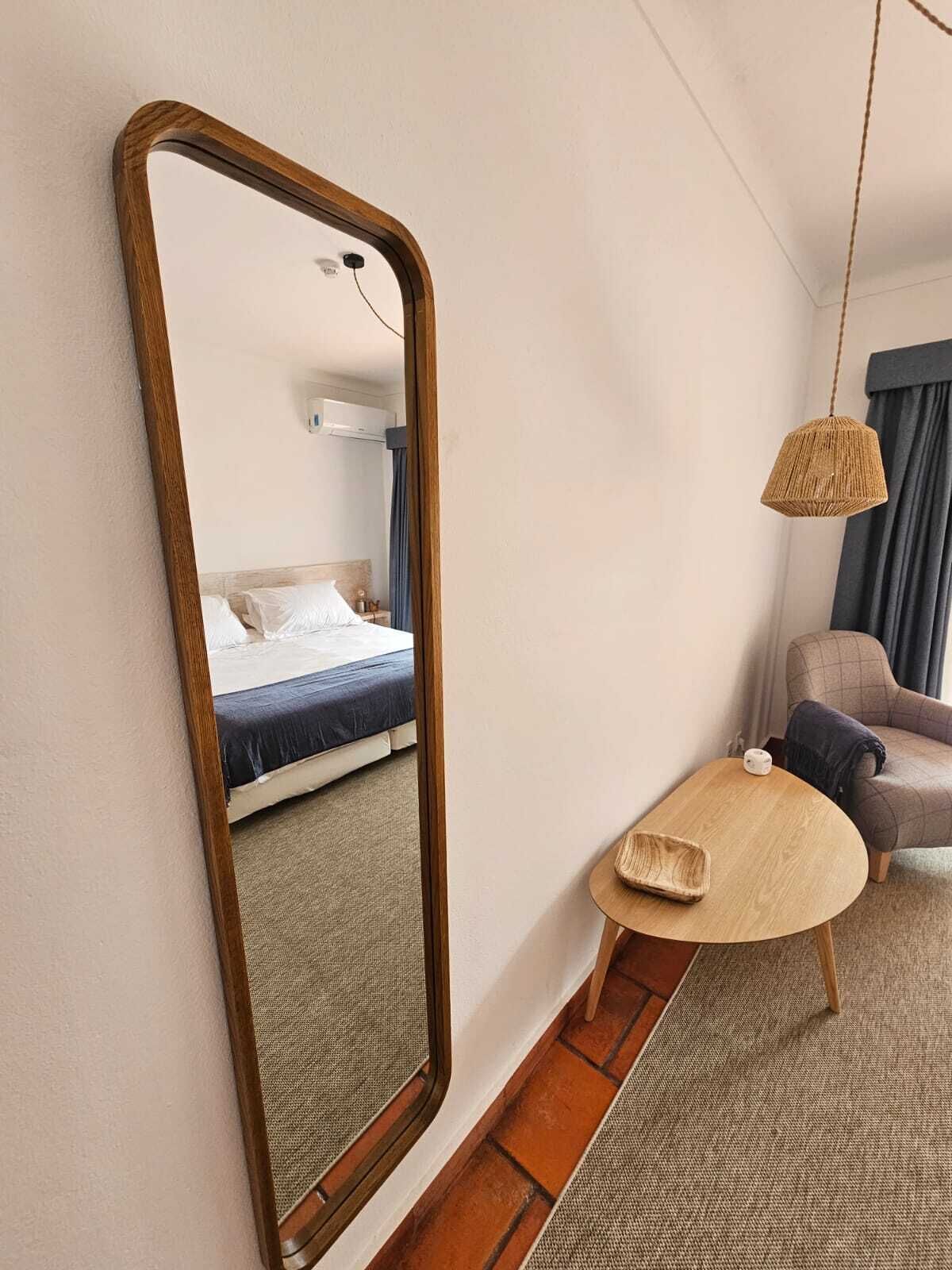 A hotel room with a large mirror, a bed with a blue mattress, a small round table, a sofa, and a hanging woven light fixture.