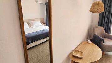 A hotel room with a large mirror, a bed with a blue mattress, a small round table, a sofa, and a hanging woven light fixture.
