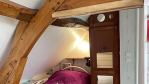 Un dormitorio pequeño y acogedor con techo inclinado, una cama individual con manta morada, una puerta de madera y una mesita con lámpara.