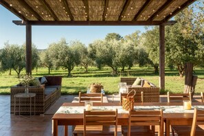 An outdoor patio with wooden furniture, a pergola with string lights, and a view of an olive orchard.