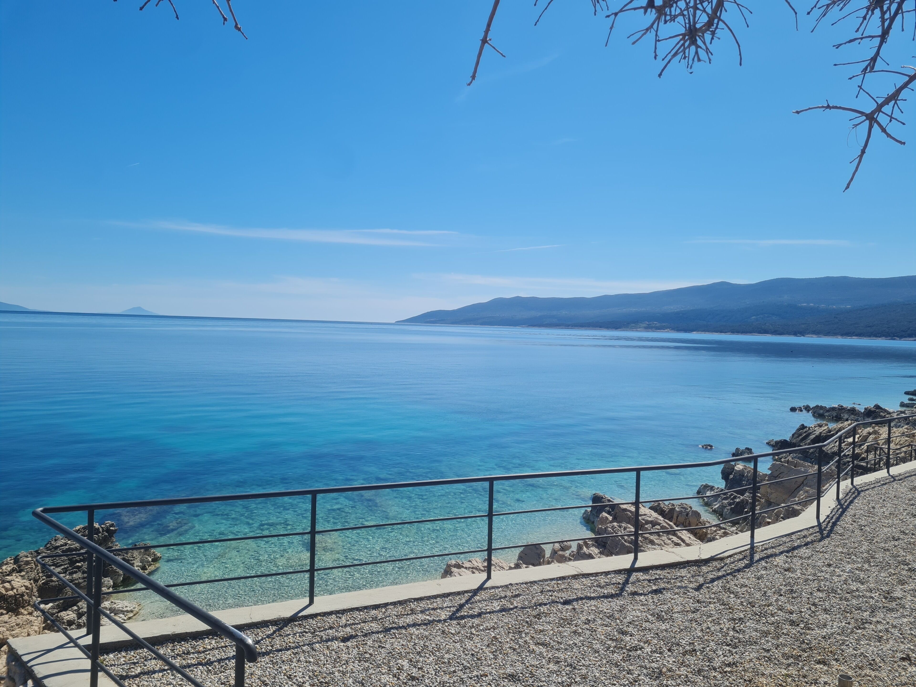 A coastal view with clear blue water, rocky shoreline, and distant mountains.