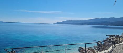 A coastal view with clear blue water, rocky shoreline, and distant mountains.
