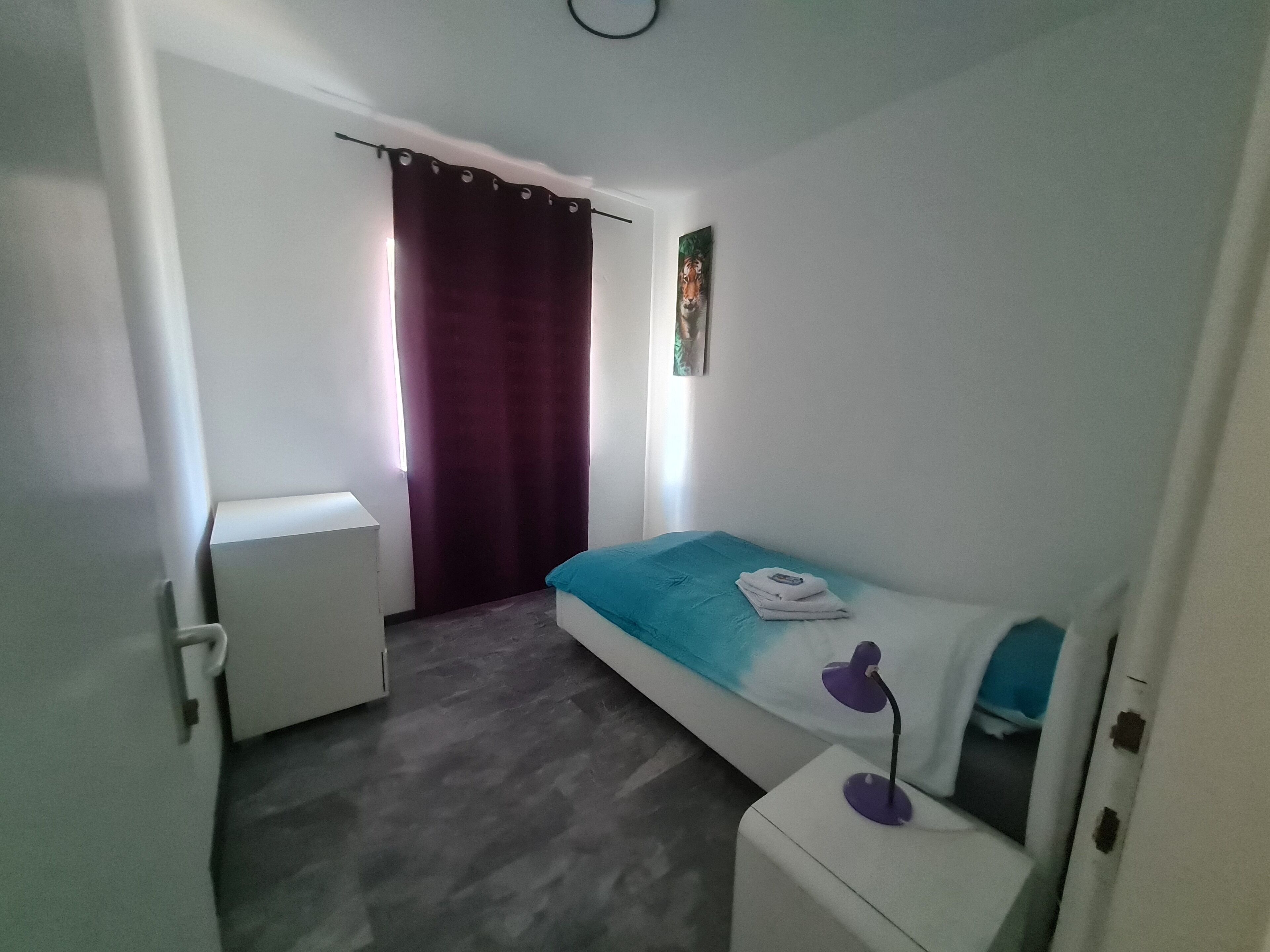 Une petite chambre à coucher, bien rangée, avec un lit, une table de chevet, une petite armoire, un rideau et un tableau accroché au mur.