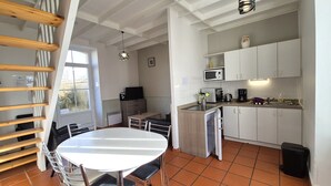 Cocina compacta con armarios blancos, microondas y un lavamanos. Hay una mesa de comedor con sillas y un ventanal que proporciona luz natural.