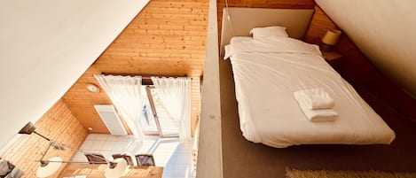 Un dormitorio con techo de madera, una cama con ropa de cama blanca, una mesita de noche pequeña y una ventana con cortinas.
