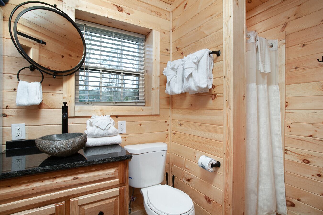 A wooden cabin bathroom with a stone basin, white toilet, and a window with blinds.