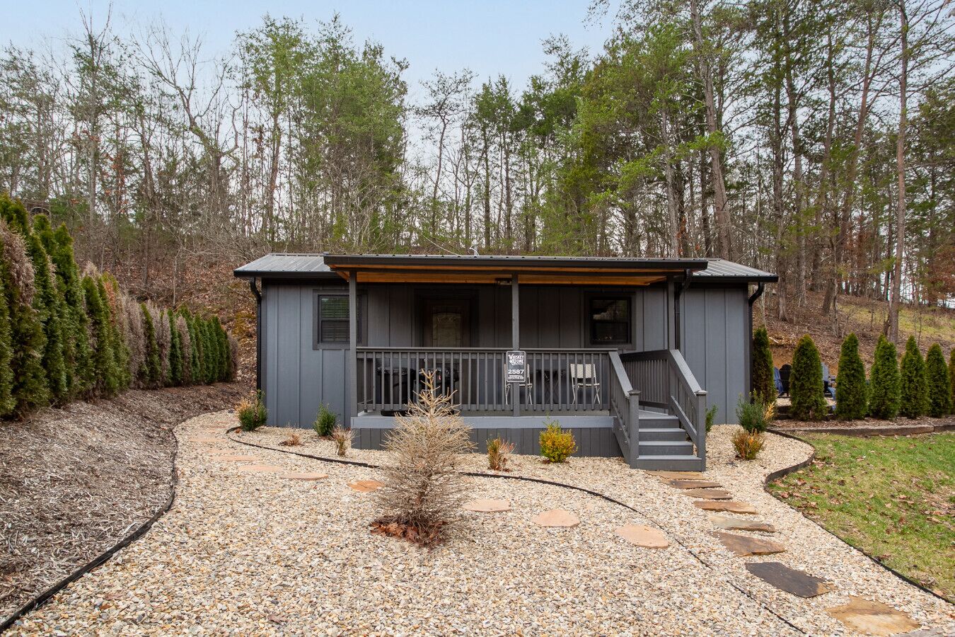 A small, single-story cabin with a porch, surrounded by a gravel driveway and a landscaped garden.