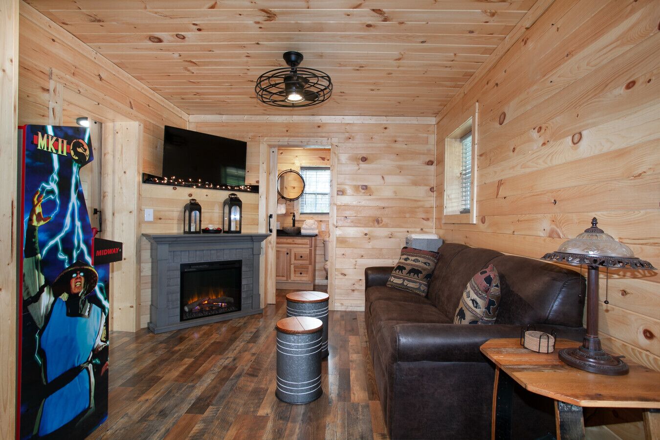 A cozy cabin interior with a fireplace, leather sofa, and a Midway arcade sign.