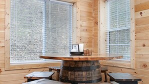 A rustic wooden interior with a round table surrounded by four stools, large windows with blinds, and a wooden floor.