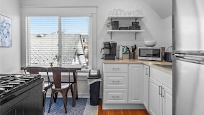 Cocina compacta con armarios blancos, estufa, microondas y refrigerador. Se aprecia una ventana con persianas, un cubo de basura y un comedor con sillas.