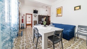 Un coin repas avec une table, des chaises et un canapé bleu. Un téléviseur est fixé au mur, et un extincteur est également accroché au mur.