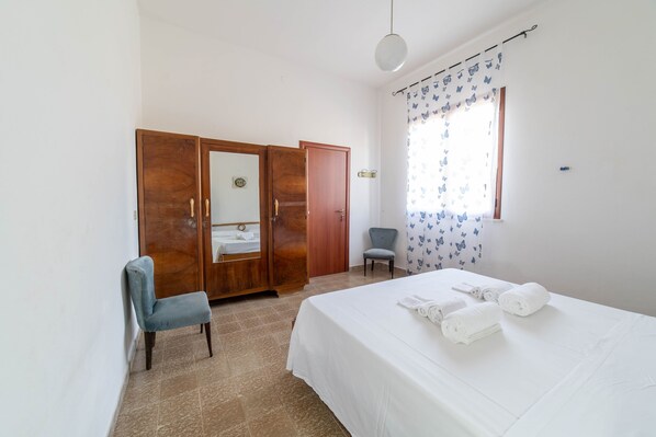Un dormitorio con cama, una silla, armario y ventana con cortinas.