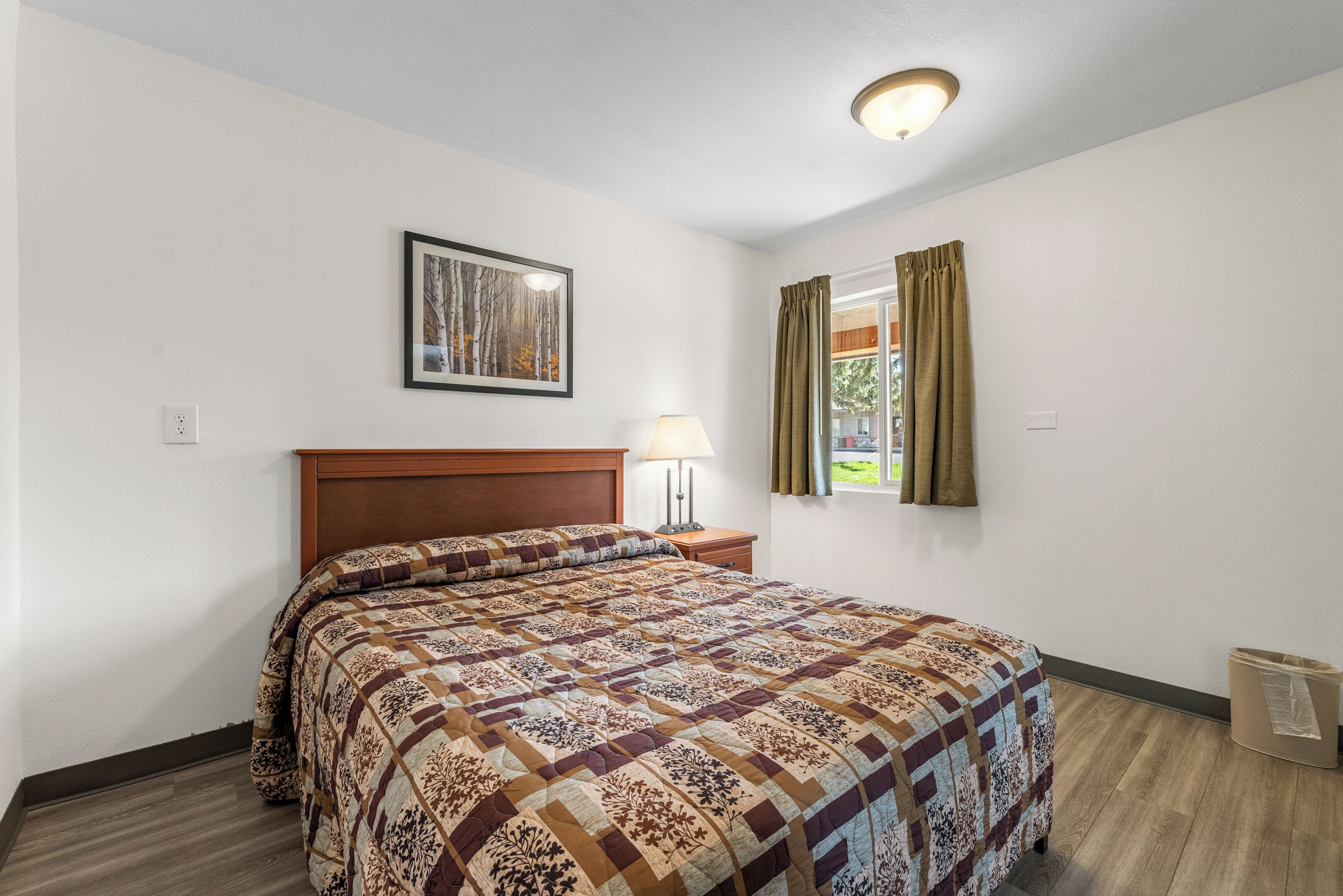 Een slaapkamer met een houten hoofdbord, een bed met een quilt met een patroon, een bedlamp, een lijst met een afbeelding aan de muur, en een raam met gordijnen.