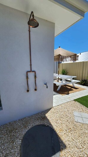 A wall-mounted outdoor shower with a hose and a faucet, a white outdoor table, and a thatched umbrella.