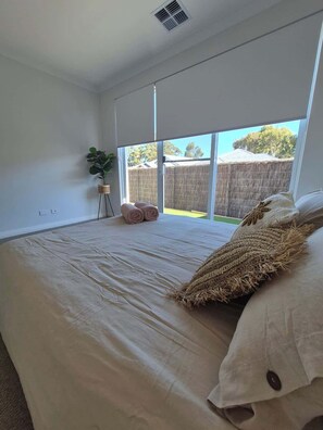 Quarto com uma cama grande, uma planta em vaso e vista para um quintal cercado.