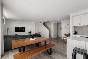 A modern living space with a wooden dining table, a sofa, a kitchen area, and a staircase.