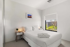 Une chambre moderne avec un grand lit, une table de chevet avec une lampe, une petite plante et une fenêtre donnant sur un paysage verdoyant.