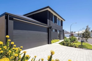A modern two-story house with a large garage, a paved driveway, and a landscaped garden with yellow flowers.