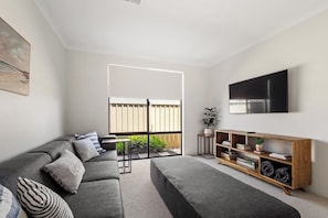 Un salon moderne avec un canapé, un pouf, un téléviseur et un meuble TV en bois.