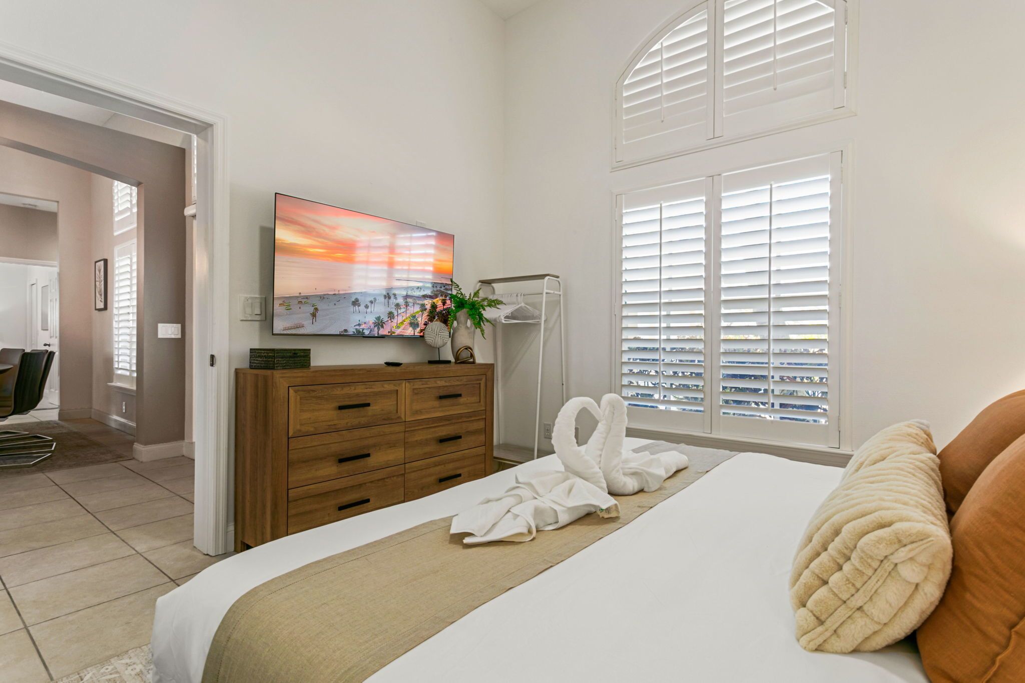 A hotel room with a bed, a TV, a wooden dresser, a plant, and a window with blinds.