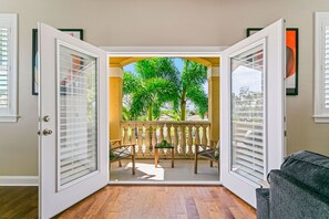 Une porte ouverte donnant sur un balcon avec une table et des chaises, une vue sur des palmiers et un canapé gris à l’intérieur.
