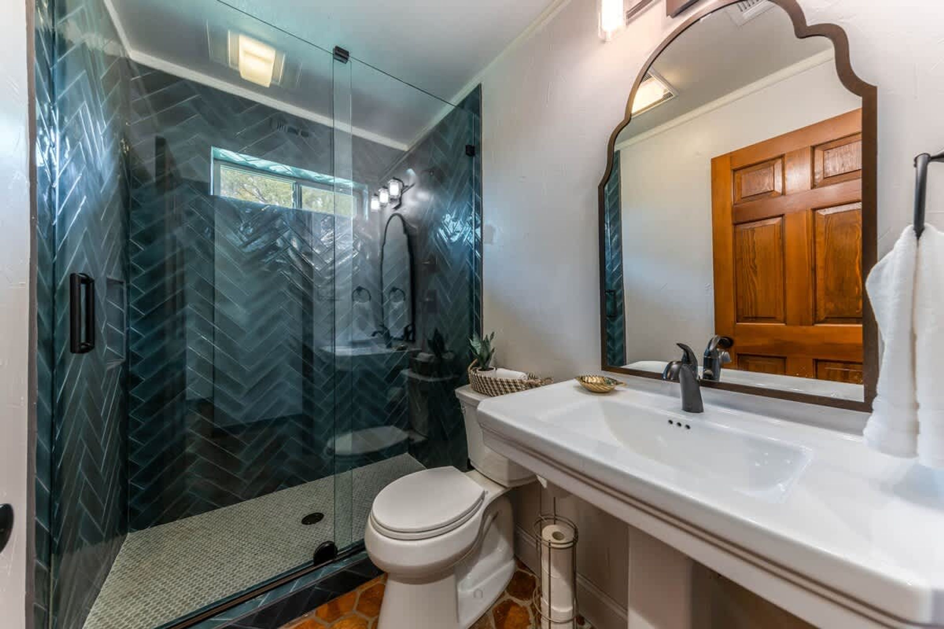 A bathroom with a glass-enclosed shower, a white toilet, a sink with a mirror, and a wooden door.