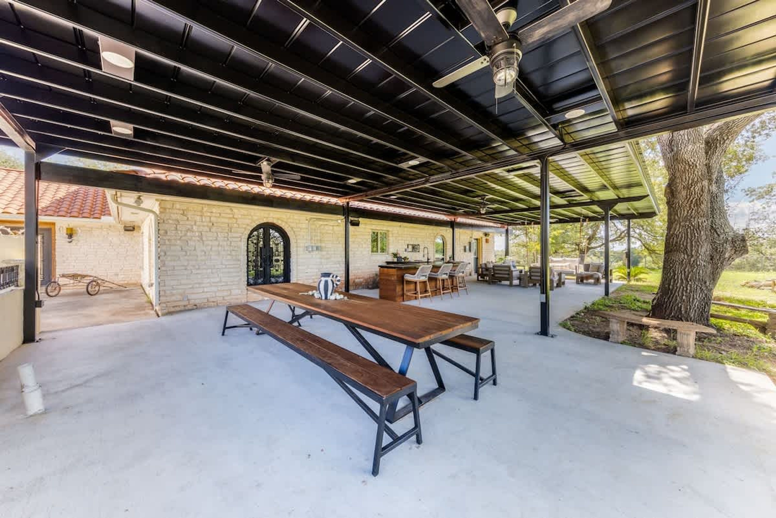 An outdoor patio with a long wooden table and benches, surrounded by chairs and a small bar area.